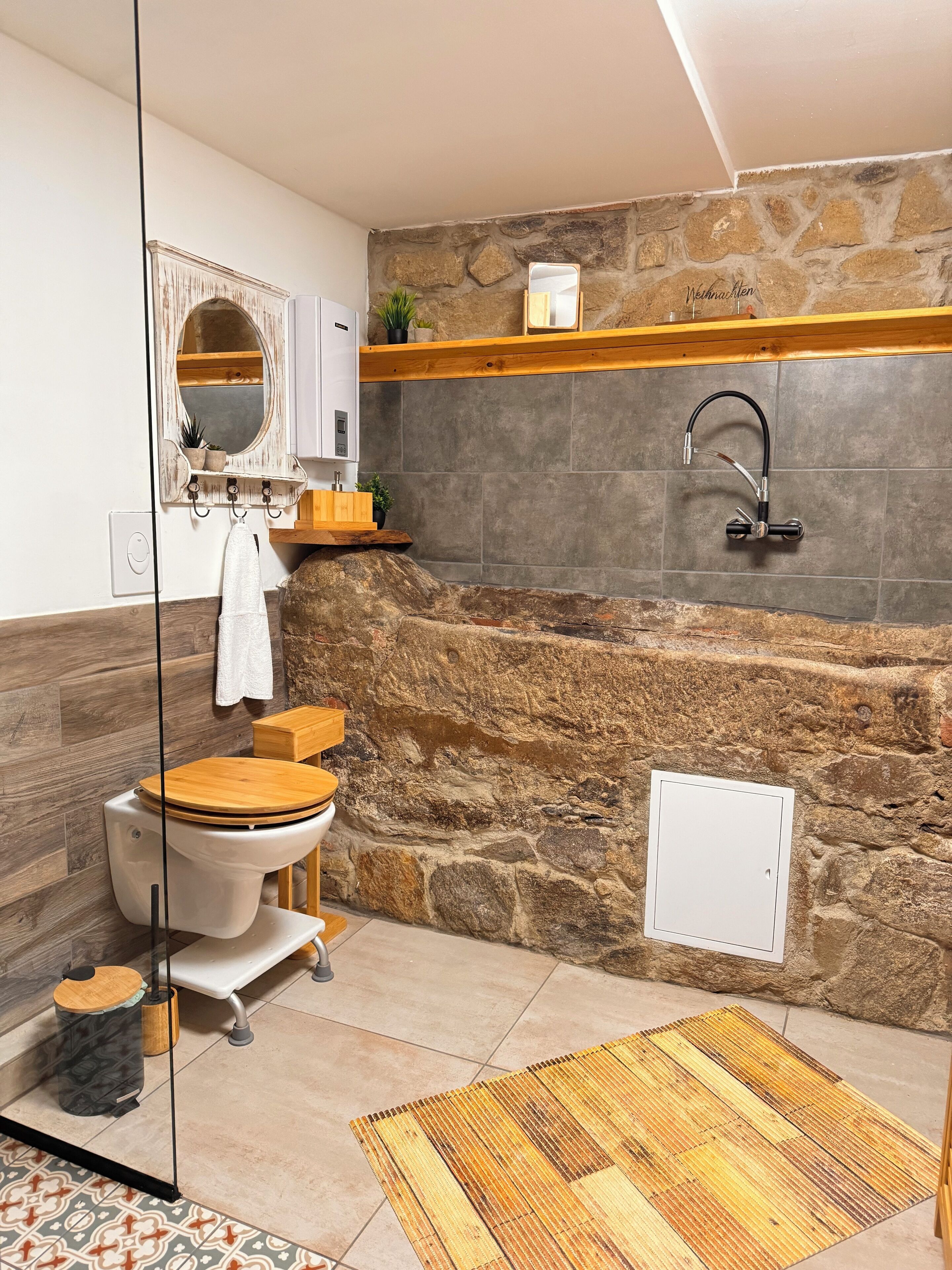 Une salle de bain avec un mur en pierre, un meuble-lavabo en bois, des toilettes, un lavabo doté d’un robinet moderne et un miroir encadré.