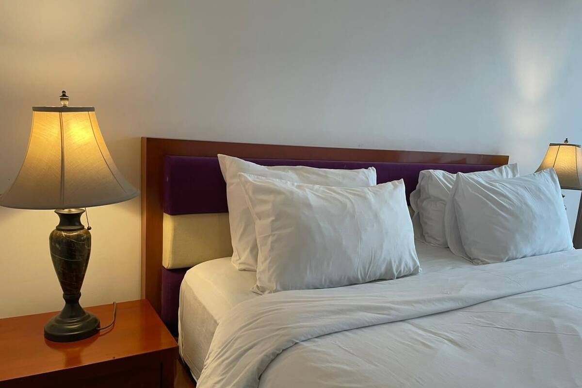 A neatly made bed with white linens and pillows, a wooden nightstand with a lamp, and a purple headboard.