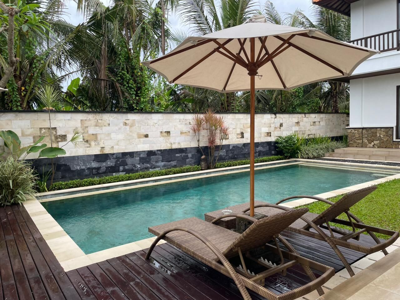 A swimming pool with lounge chairs and an umbrella, surrounded by a stone wall and tropical plants.