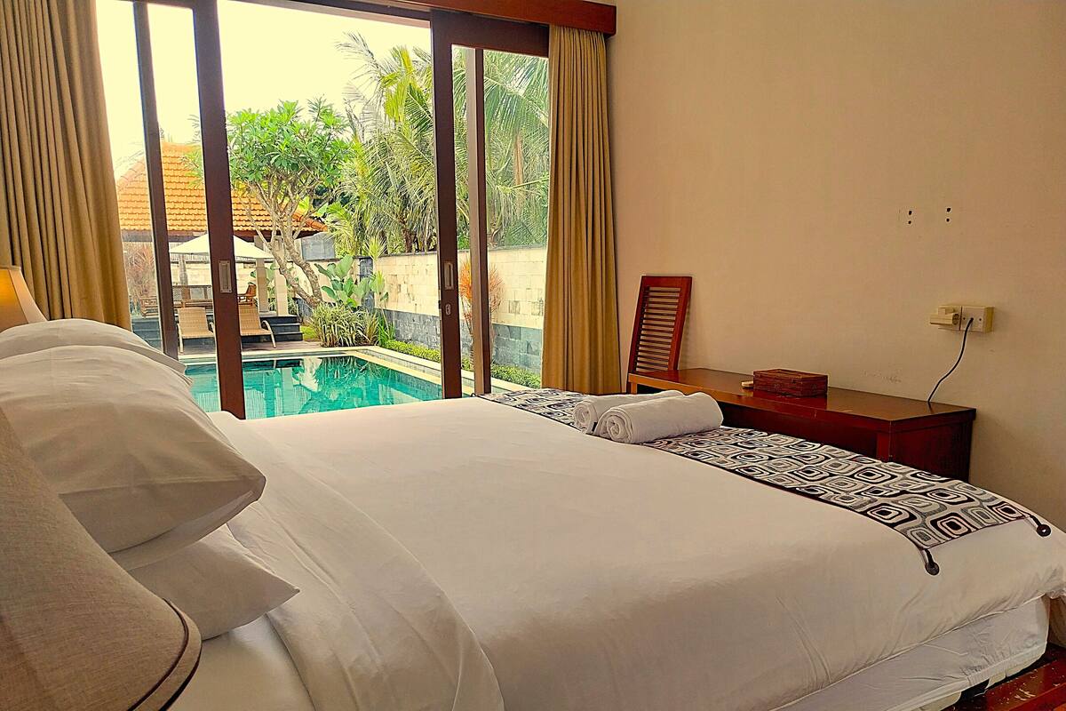 A hotel room with a large bed, a small desk, a chair, and a view of an outdoor swimming pool.