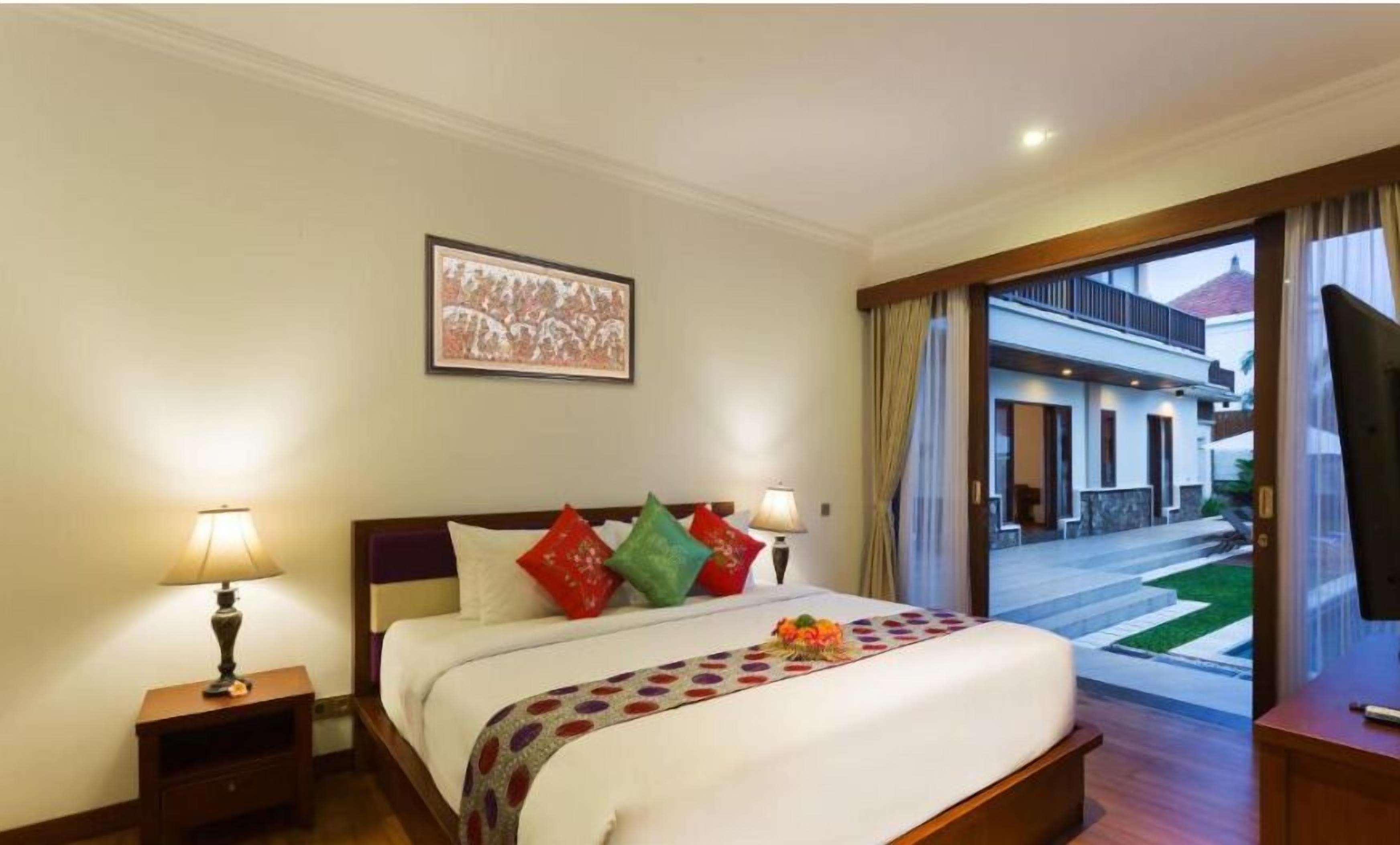 A hotel room with a bed, two bedside lamps, a framed picture on the wall, and a view of the outside through a glass door.
