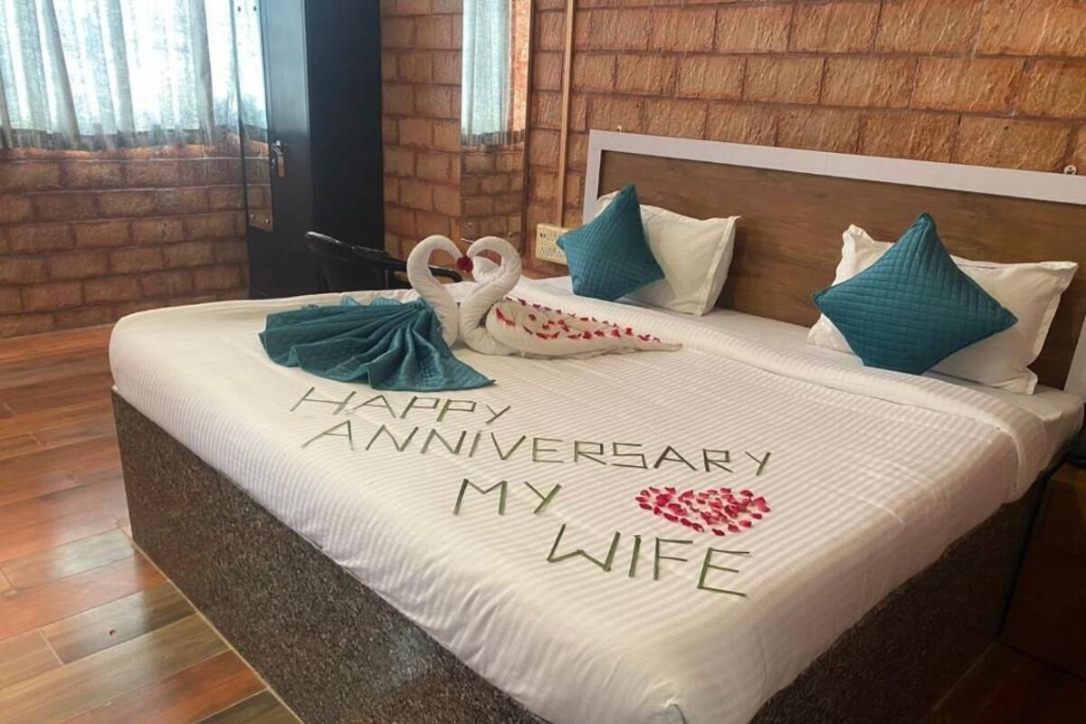 A hotel room with a bed featuring a 'Happy Anniversary My Wife' bedspread, a wooden headboard, and a brick wall.