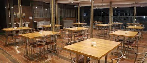 A restaurant with multiple wooden tables and metal chairs, large windows, and a kitchen area visible in the background.