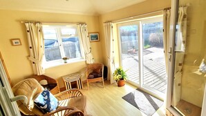 A room with a wooden floor, a wicker chair, a small table, a potted plant, and a sliding glass door leading to an outdoor deck. - Spacious holiday home with wrap around gardens (Perranporth)