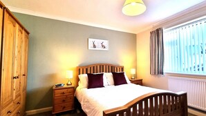 A bedroom with a wooden bed, two bedside tables with lamps, a wardrobe, and a window with blinds. - Spacious holiday home with wrap around gardens (Perranporth)