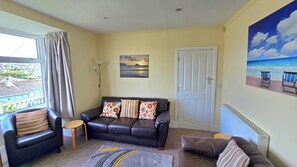 A living room with a sofa, a small table, a painting of a sunset, and a view of the sea through a window.