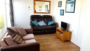 A living room with a brown sofa, a black leather couch, and a wooden TV stand. - Chalet on Perran Sands, Haven holiday park (Perranporth)