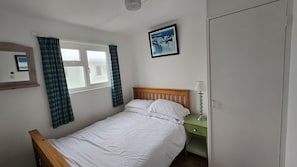 A bedroom with a bed, a bedside table, a lamp, a window with curtains, a framed picture on the wall, and a wardrobe.