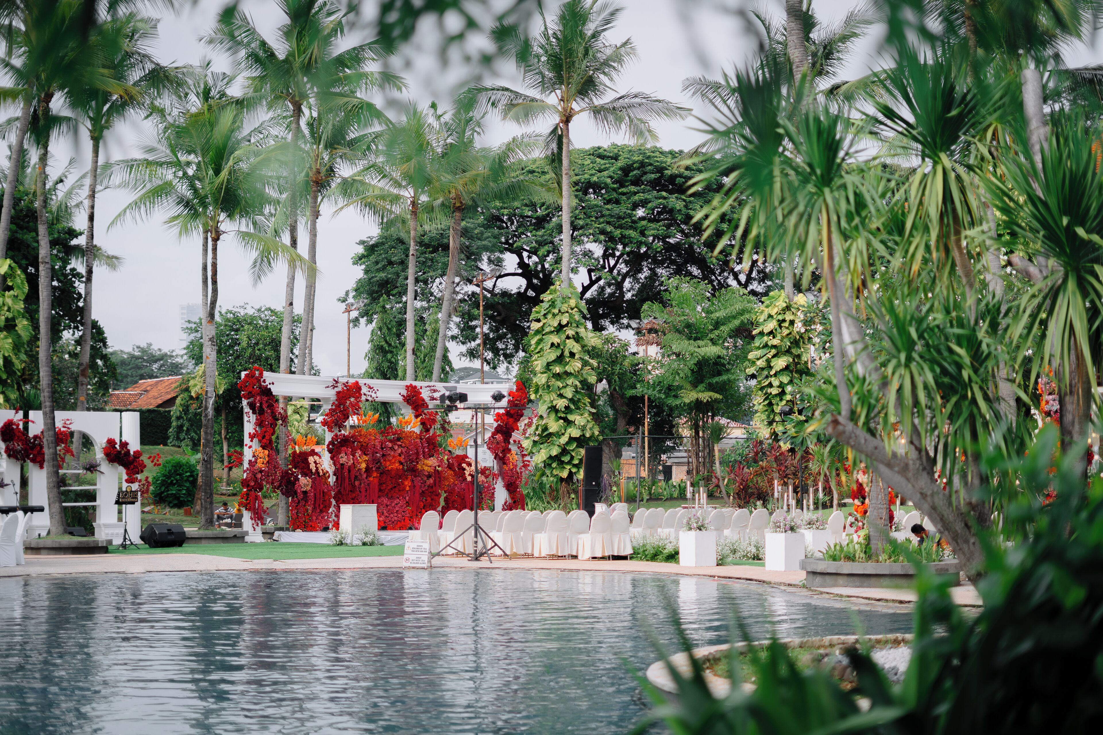 A poolside area with a stage decorated with red flowers, surrounded by palm trees and a white fence.