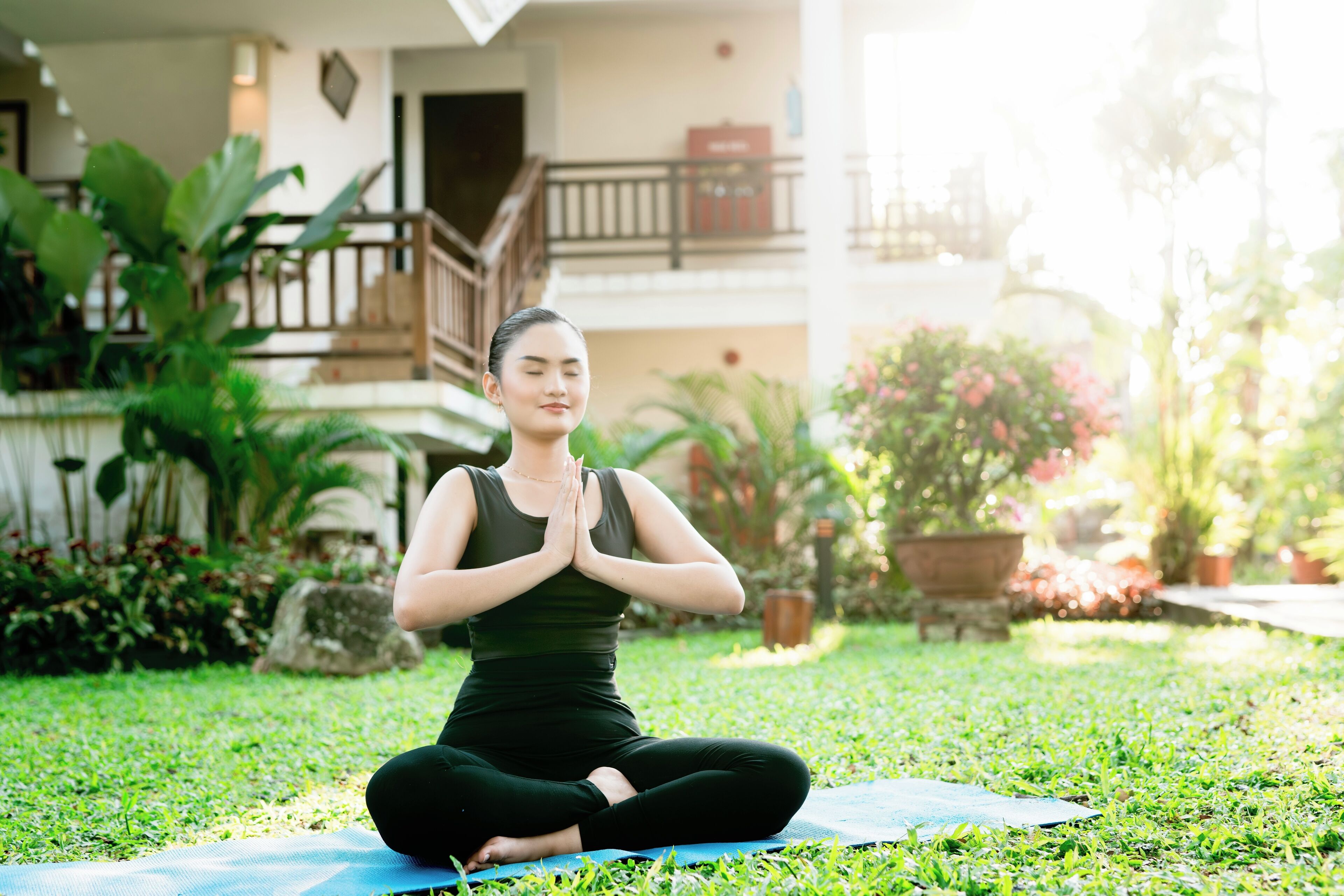 A person sitting on a yoga mat in a garden area with a building in the background.