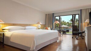 Une chambre d’hôtel avec un grand lit, un bureau avec une chaise, un ordinateur portable et une vue sur les palmiers par la fenêtre.