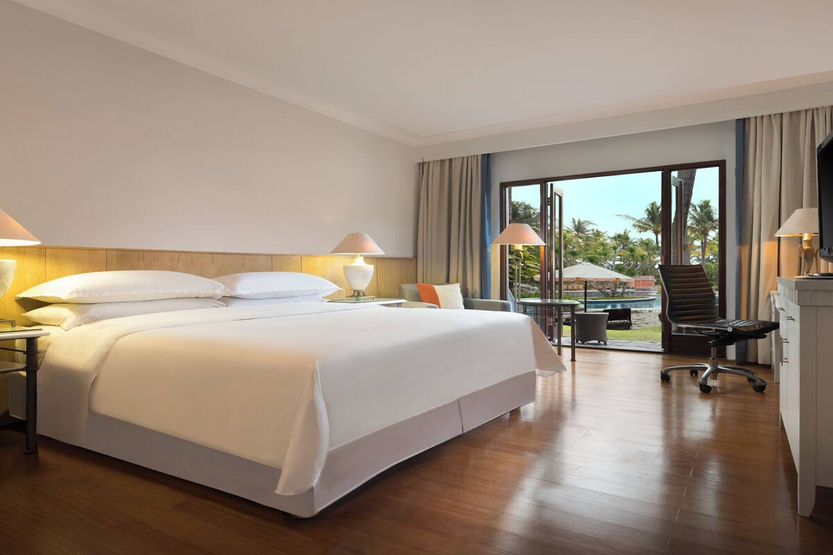 Habitación de hotel con una cama grande, un escritorio con silla, una computadora portátil y vistas a palmeras a través del ventanal.