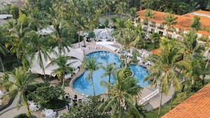 Un complexe hôtelier doté d’une grande piscine centrale entourée de palmiers, d’espaces de détente en plein air et d’un bâtiment surmonté d’un toit en tuiles.