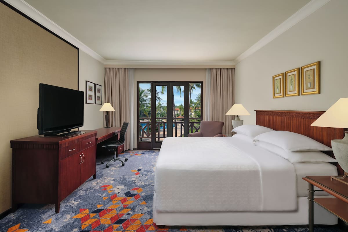 Habitación de hotel con una cama grande, un escritorio con televisor, una silla y un balcón con vistas a palmeras.