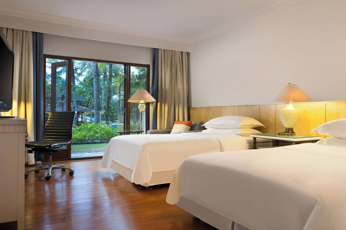 Habitación de hotel con dos camas, un escritorio, una silla y una ventana con vista a palmeras.