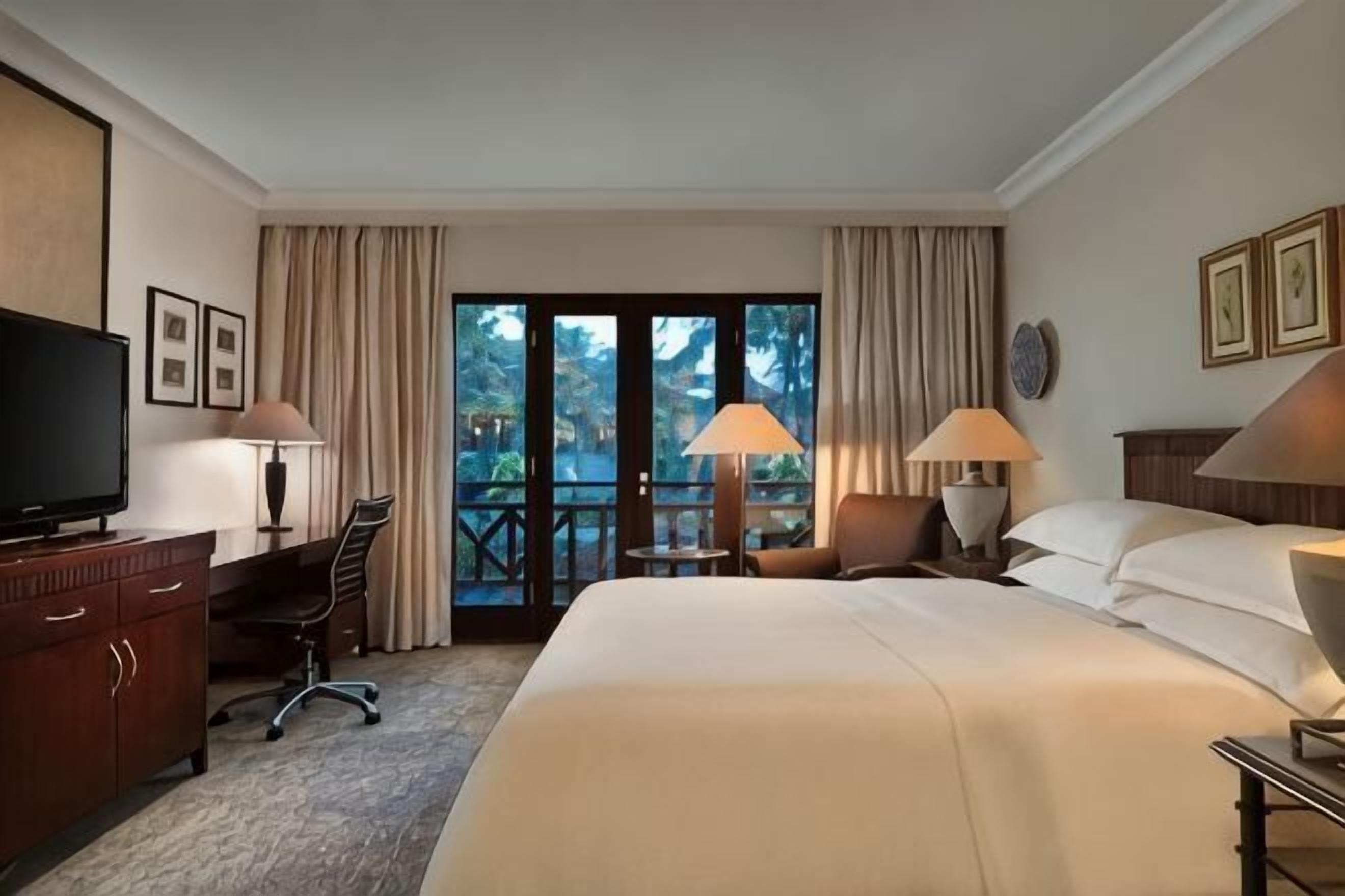 Une chambre d’hôtel équipée d’un lit, d’un bureau, d’une chaise, d’une télévision, de lampes et d’un balcon avec des rideaux.