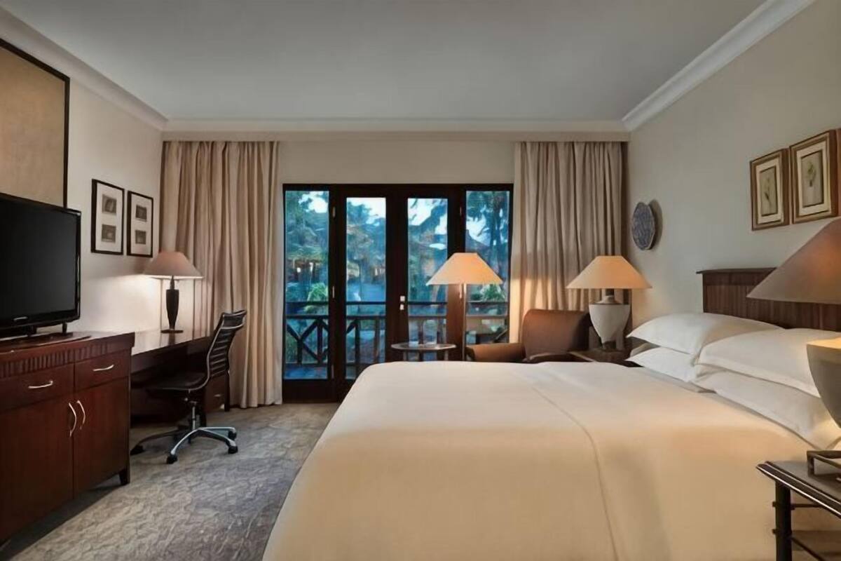 Habitación de hotel con cama, escritorio, silla, televisor, lámparas y balcón con cortinas.