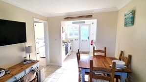 Un coin repas avec une table et des chaises en bois, un téléviseur accroché au mur et une cuisine visible à travers une porte ouverte.