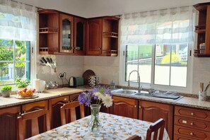 A kitchen with wooden cabinets, a dining table with a floral tablecloth, a vase with flowers, and a window with a view of greenery. - Casa Brisa - by PrimeStayMadeira (São Vicente)
