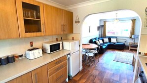 A compact kitchen with wooden cabinets, a microwave, and a toaster. There is a dining area with a round table and chairs, and a blue sofa. A sign on the wall reads "BEACH HOUSE RULES." - Beachfront apartment with direct beach access! (Perranporth)