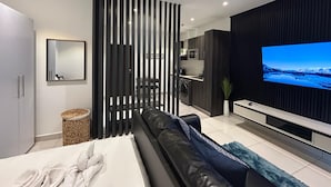A modern living room with a black sofa, a TV, and a kitchen in the background. - Embassy Gardens Haven (Accra)