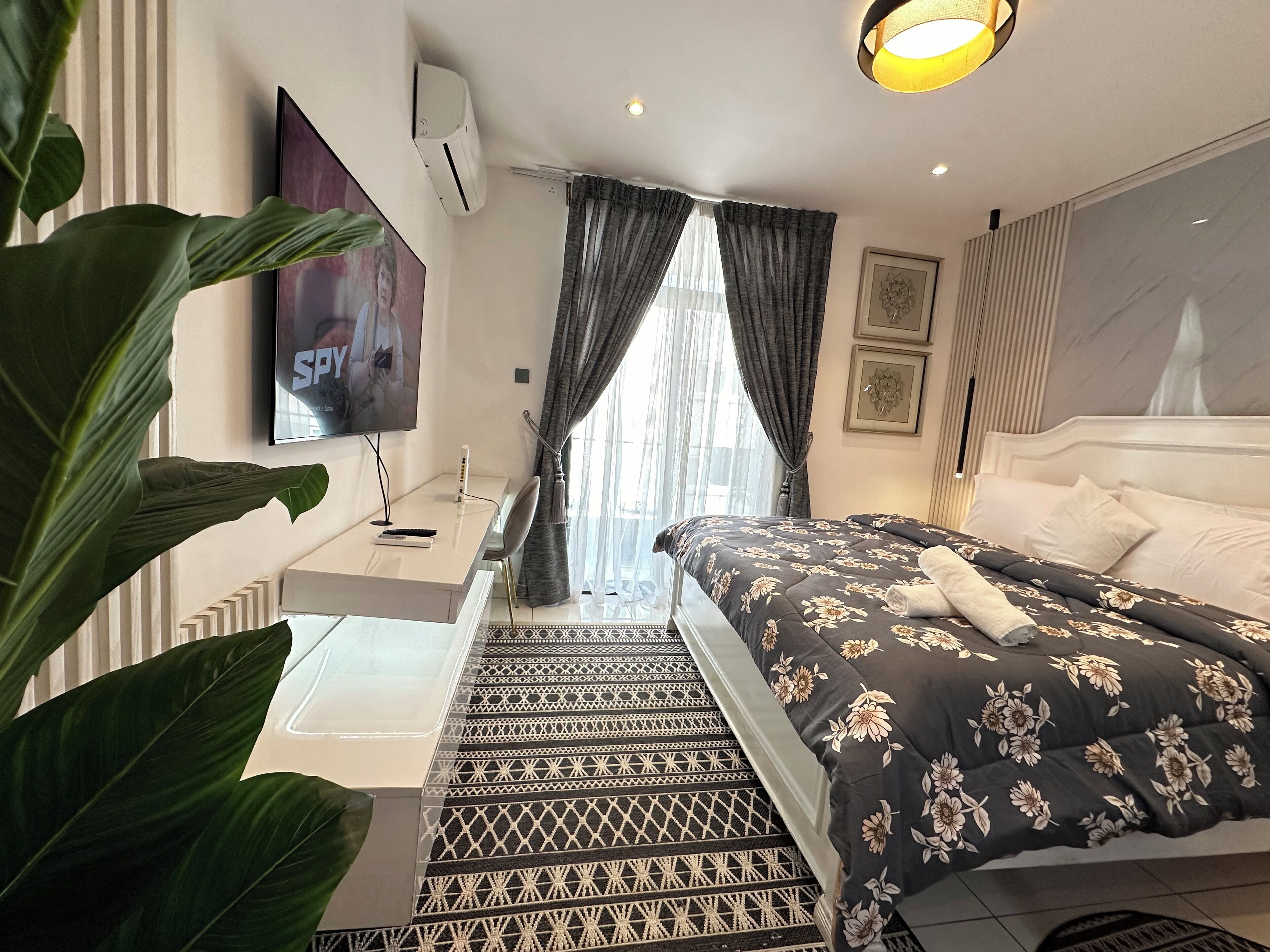A modern hotel room with a bed, a TV, a desk, and a large plant.