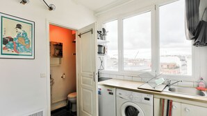 A compact kitchen with a washing machine, a sink, and a window with a view of the city. - Studio overlooking the rooftops of Paris, 2 minutes from the metro (Paris)