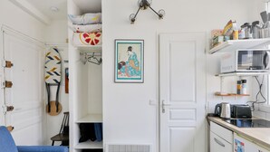 A compact kitchen with white cabinets, a microwave, and a toaster. A blue chair is visible in the foreground. - Studio overlooking the rooftops of Paris, 2 minutes from the metro (Paris)