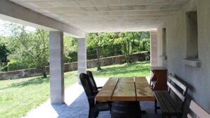 An outdoor patio with a wooden table and benches, surrounded by greenery and a stone wall.