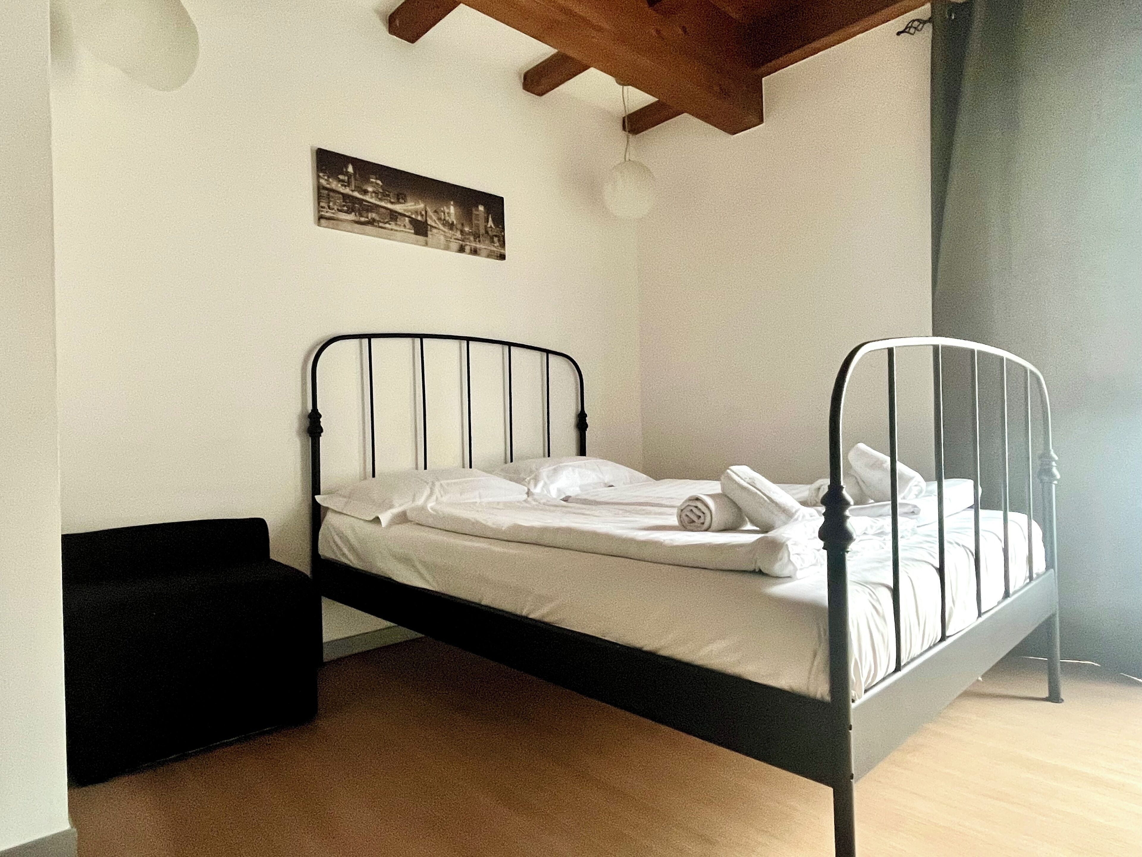 Una cama individual con estructura metálica, ropa de cama blanca y un pequeño banco negro.