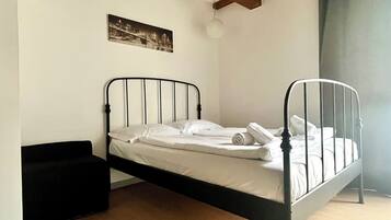 Una cama individual con estructura metálica, ropa de cama blanca y un pequeño banco negro.