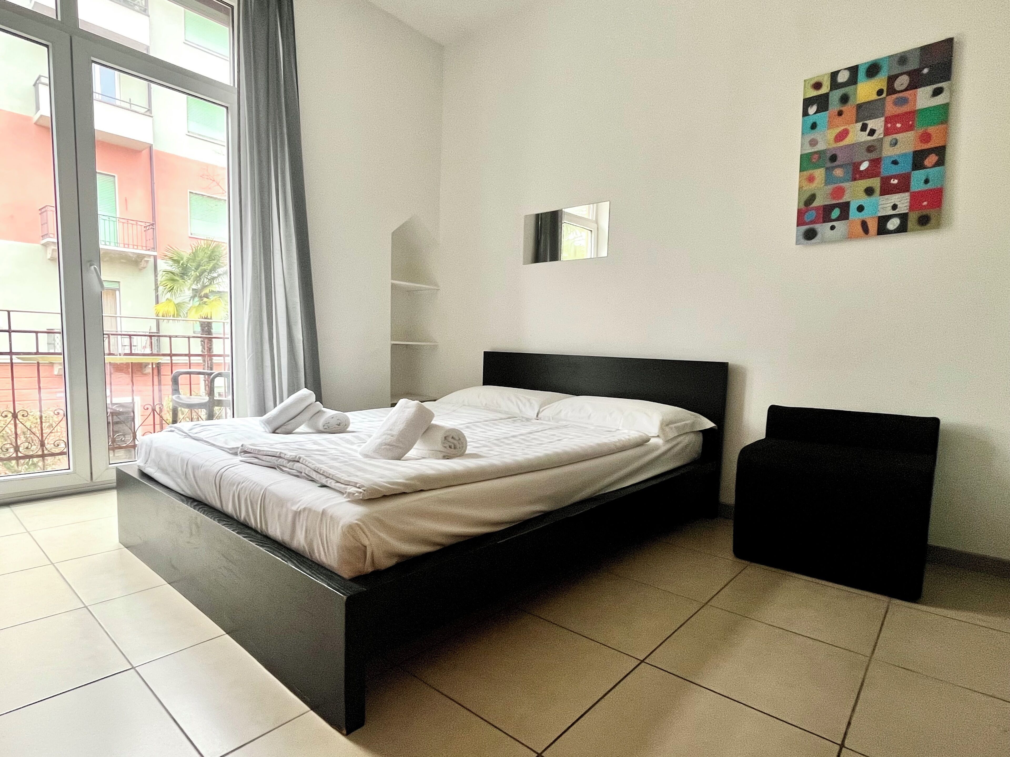 A modern hotel room with a large bed, a window with a view of buildings, and a colorful artwork on the wall.