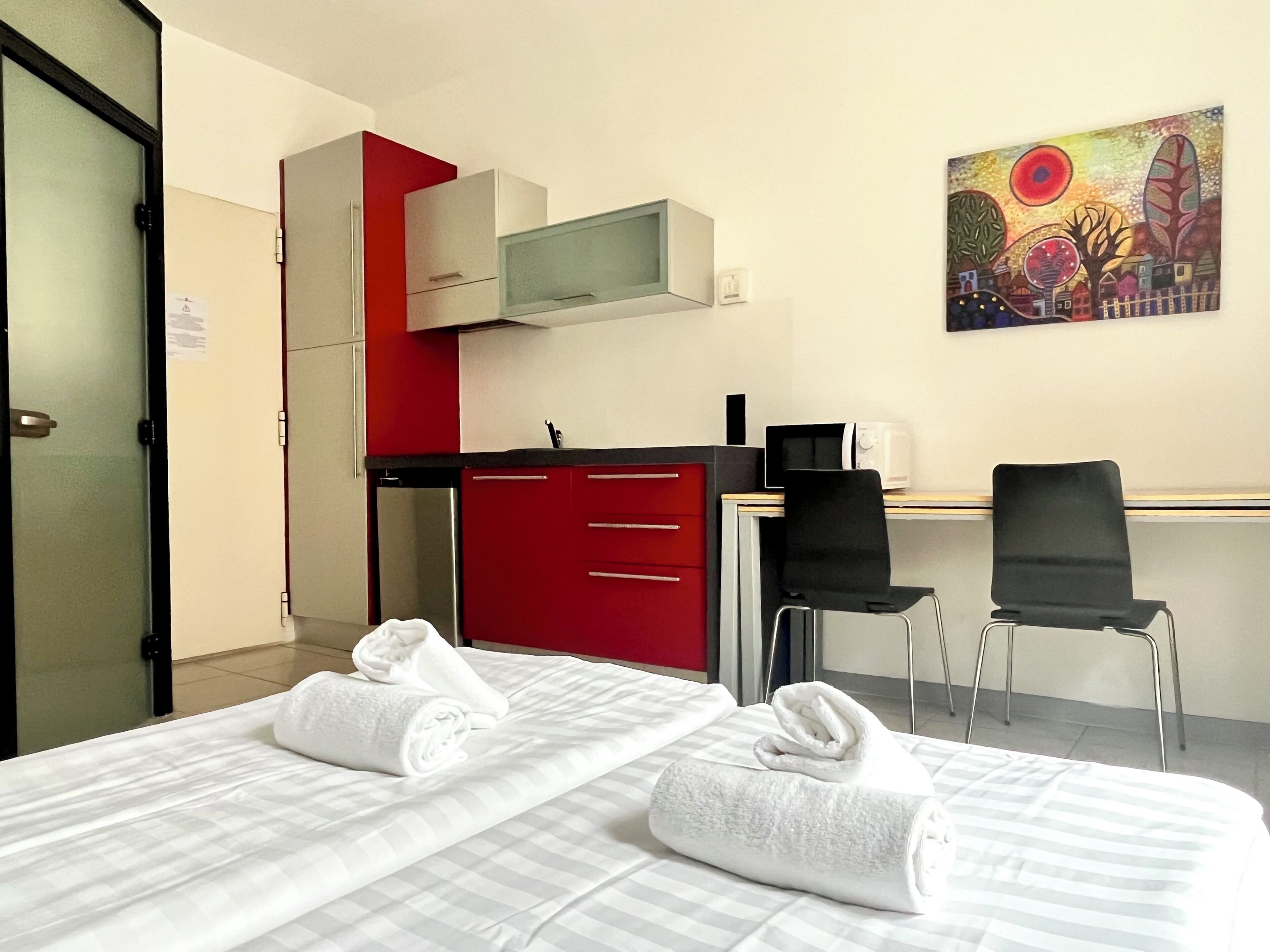 Una camera d'albergo con due letti, una scrivania con una sedia, un armadietto rosso e un quadro appeso al muro.