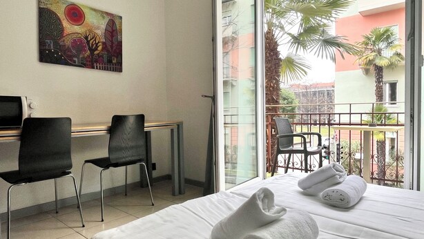A hotel room with a bed, two chairs, a desk, a painting on the wall, and a balcony with a view of palm trees and buildings. - GuestHouse Lugano University (Lugano)
