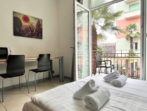 A hotel room with a bed, two chairs, a desk, a painting on the wall, and a balcony with a view of palm trees and buildings. - GuestHouse Lugano University (Lugano)