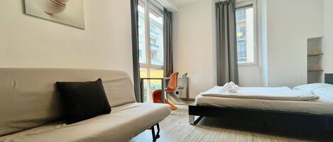 A modern hotel room with a sofa, a bed, a chair, and a large window.