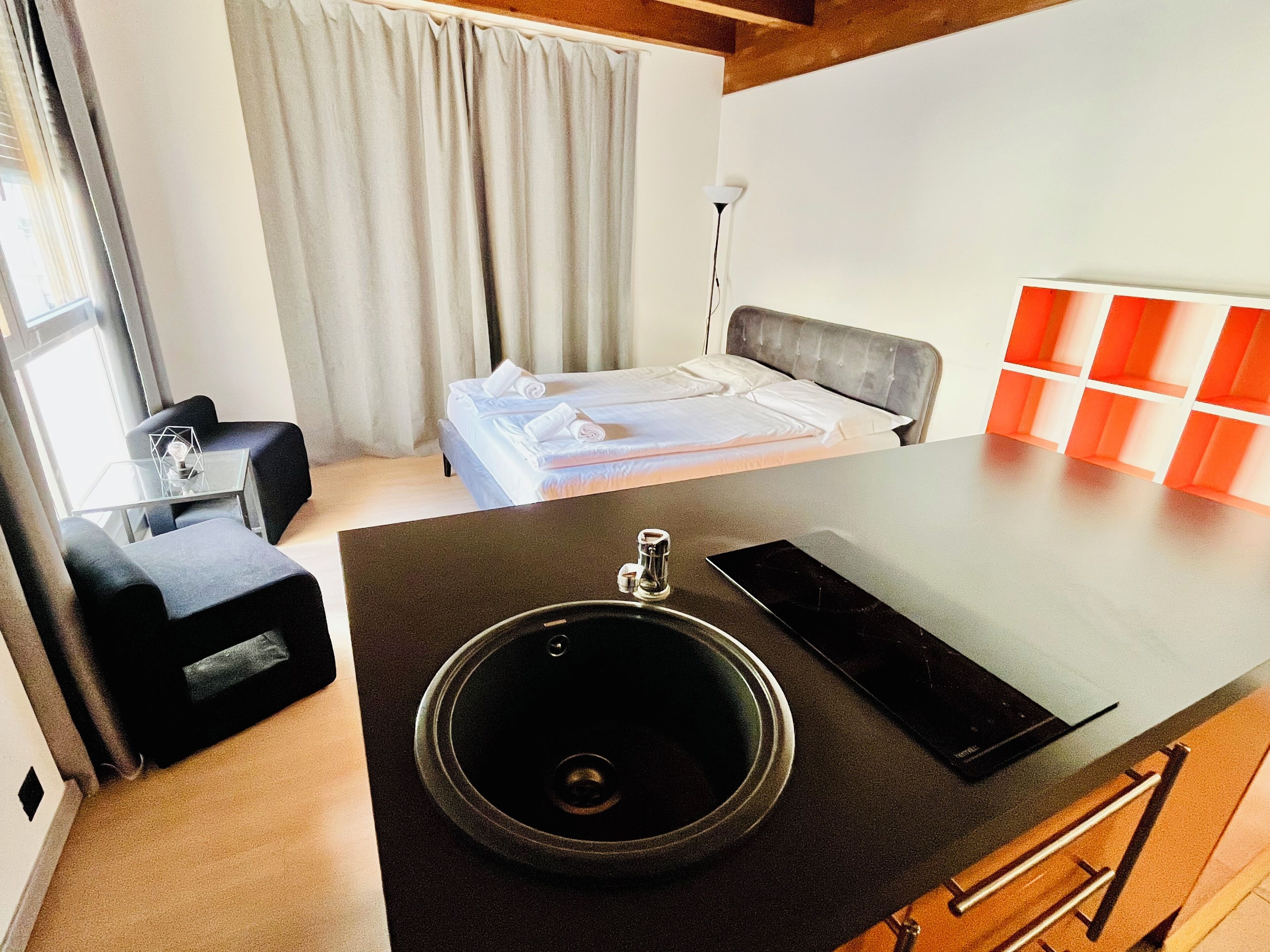 A modern hotel room with a bed, a desk, a chair, a black sink, and a small table.