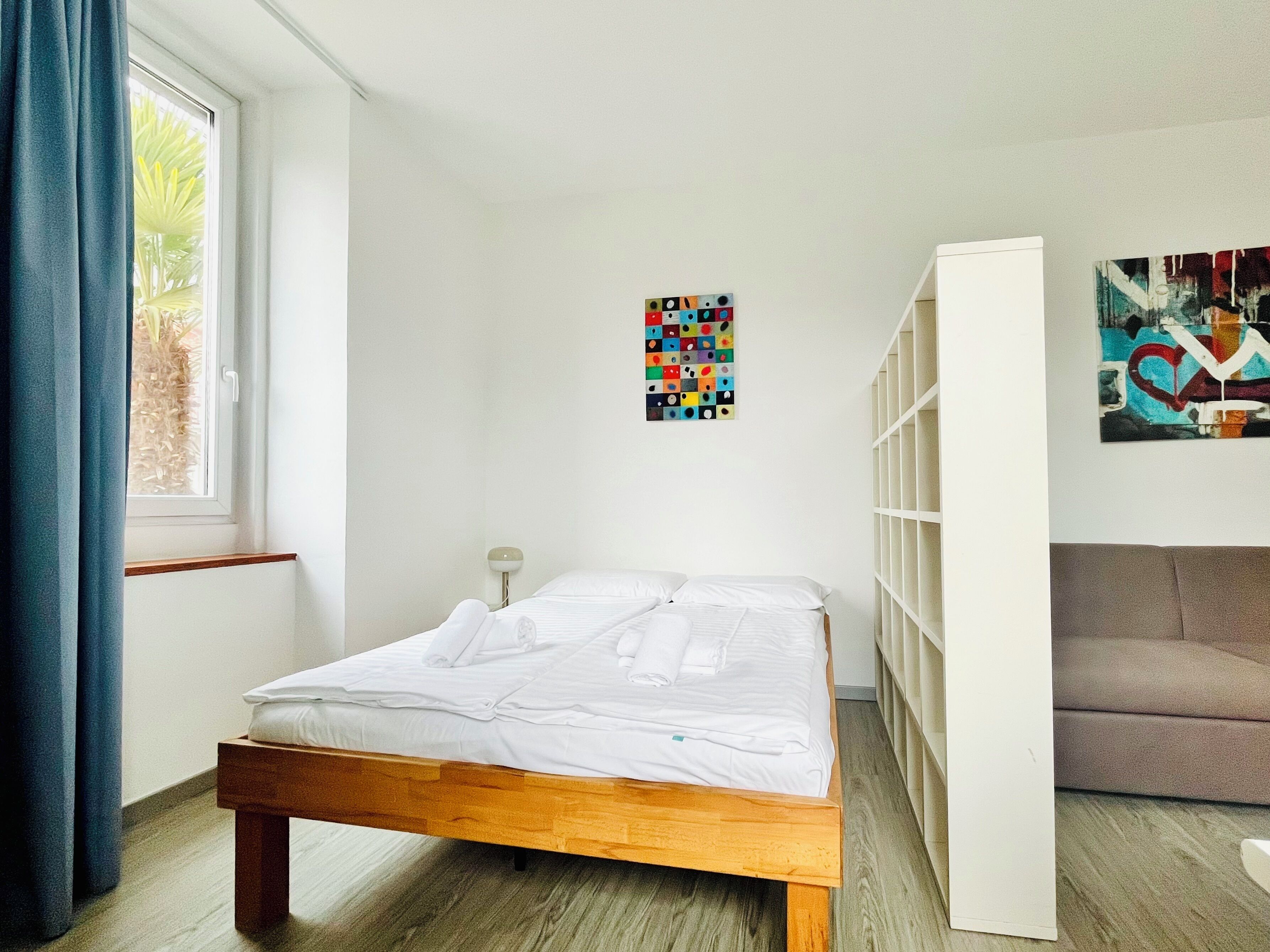 A bedroom with a wooden bed, a window with blue curtains, a wall-mounted shelf, and a colorful abstract painting.