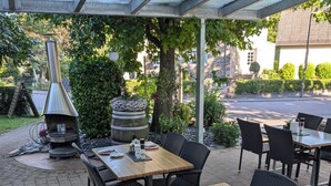 Una terrazza esterna con tavoli in legno e sedie in vimini nero, una botte in pietra e un camino a stufa in metallo.