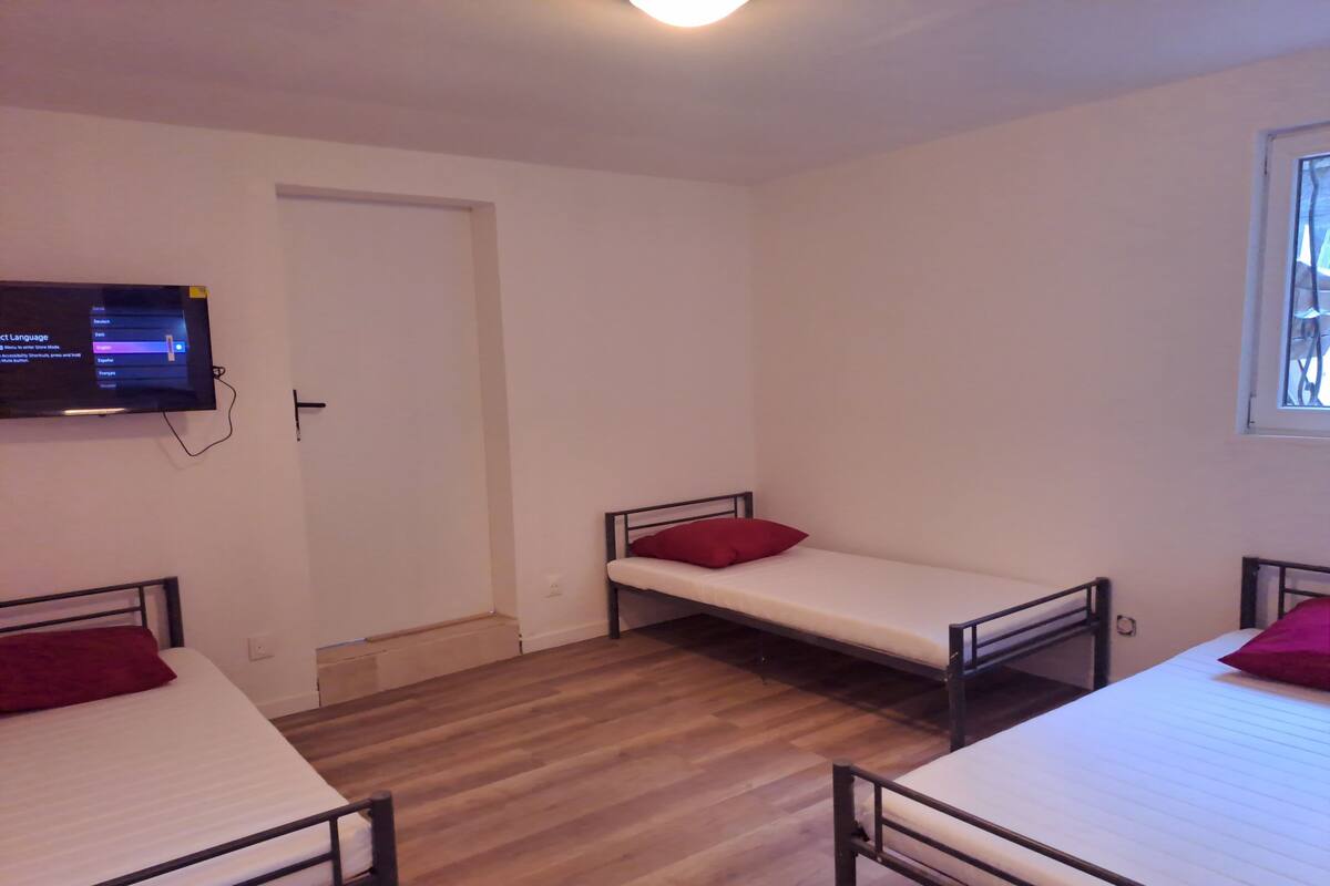 Une chambre avec trois lits simples, chacun agrémenté d’un oreiller rouge. Un téléviseur est fixé au mur, et il y a une fenêtre avec des barreaux.