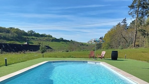 Una piscina amb un pont de fusta al primer pla, un àrea de pícnic amb menjar i begudes, i una vista panoràmica de turons i arbres al fons.