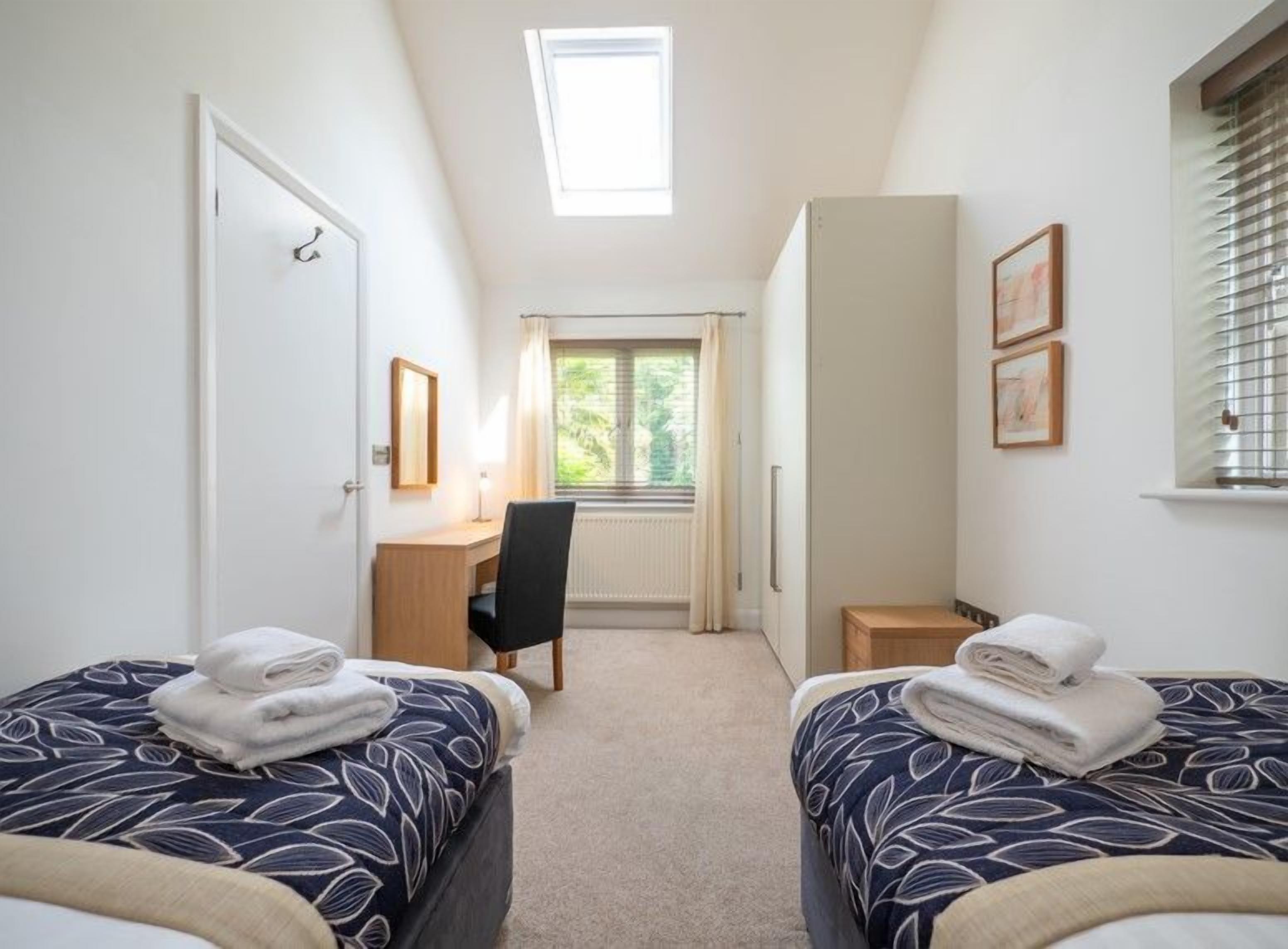 A bedroom with two beds, a desk, a chair, and a window with blinds.