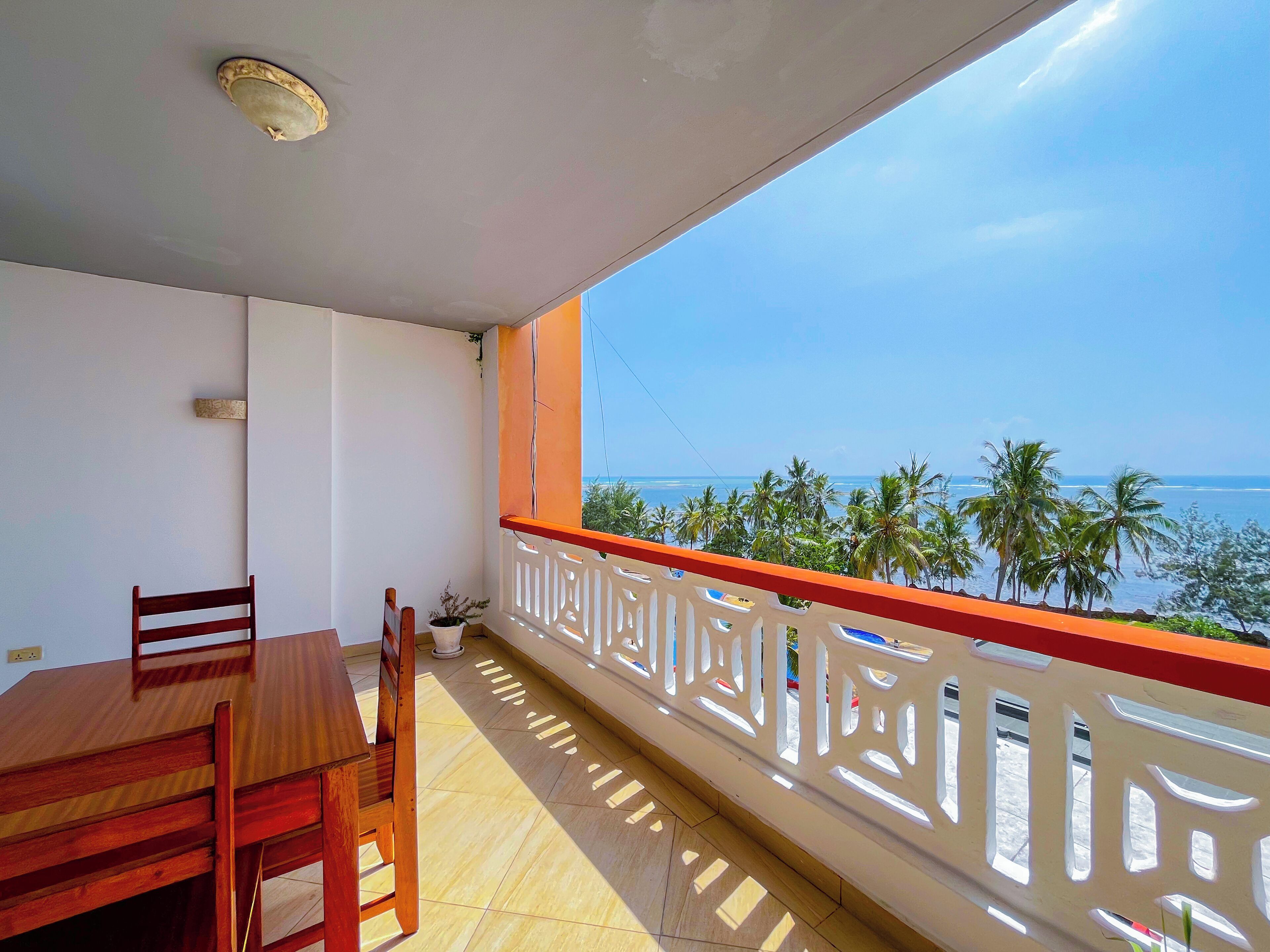 A balcony with a wooden table and chairs, a white railing, and a view of the ocean and palm trees.