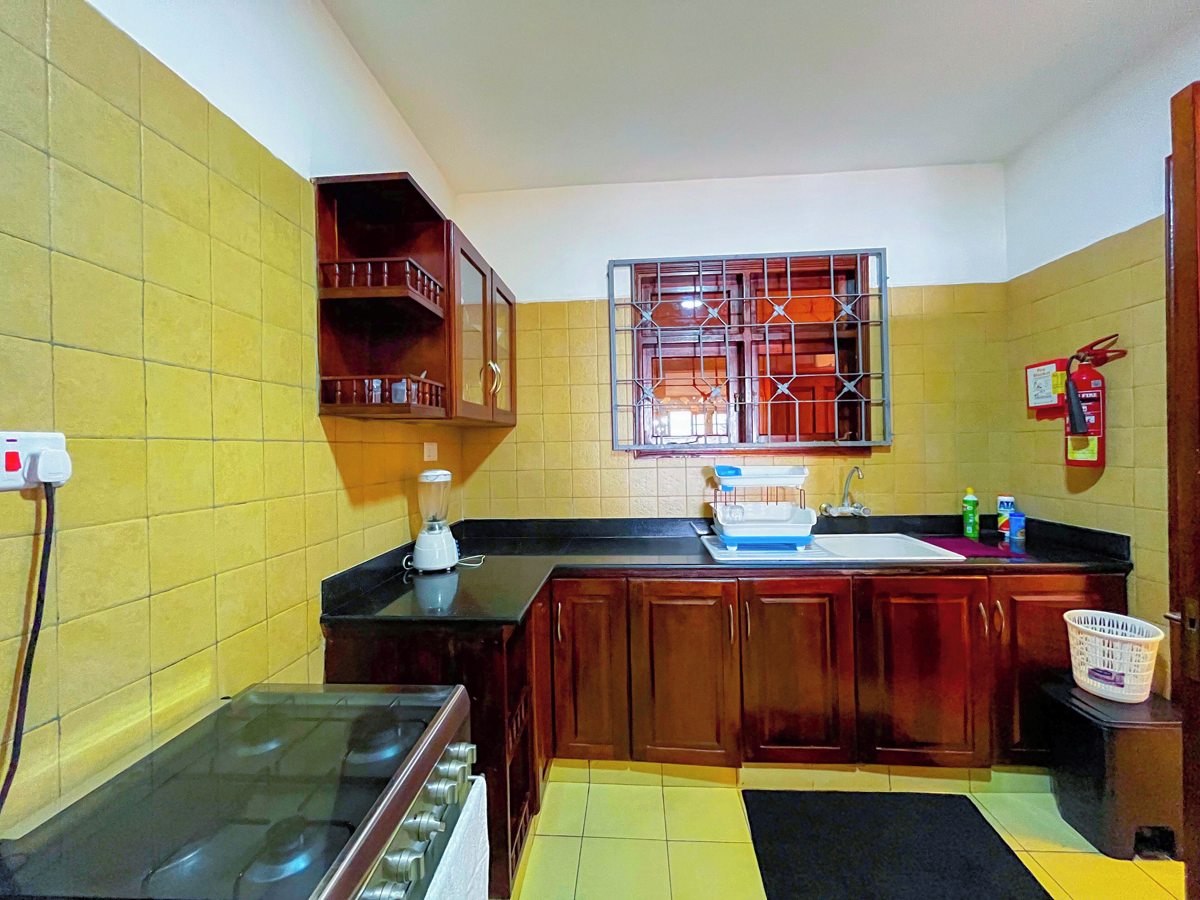 A kitchen with yellow walls, wooden cabinets, a stove, a blender, a sink, and a window with bars.