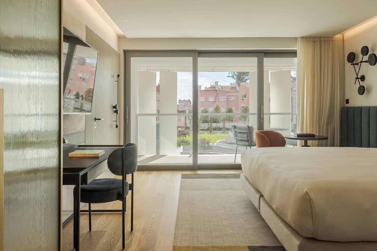 A modern hotel room with a large bed, a desk with a chair, a small table, and a view of the city through large windows.