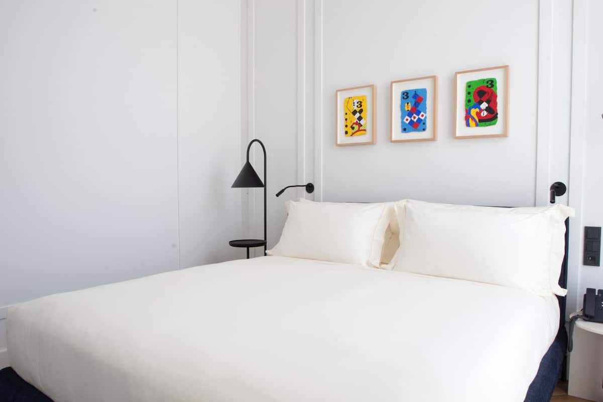 A modern bedroom with a large bed, a bedside table, a lamp, and three framed artworks on the wall.