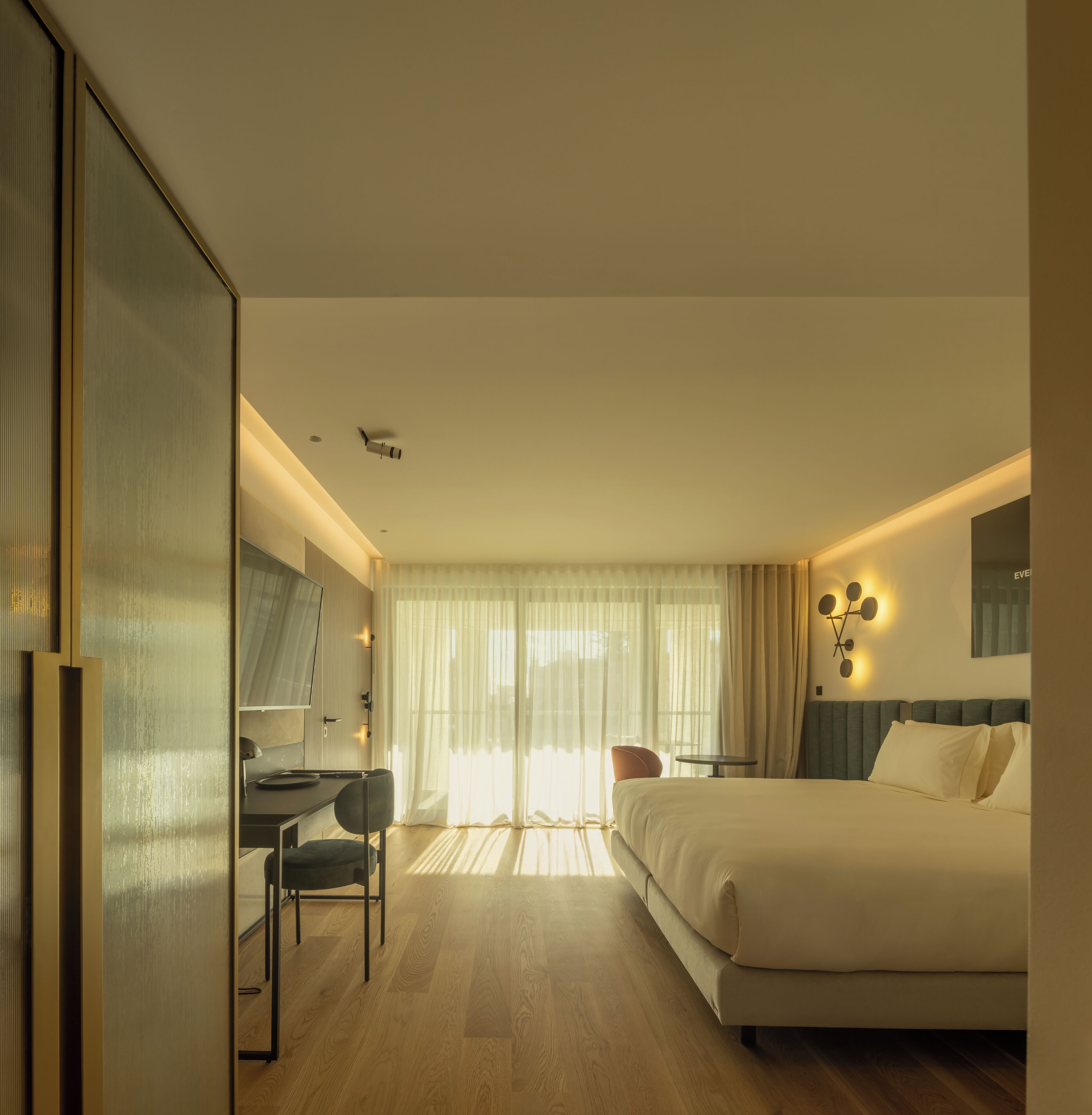 A modern hotel room with a large bed, a desk with a chair, a small table, and a mirror.