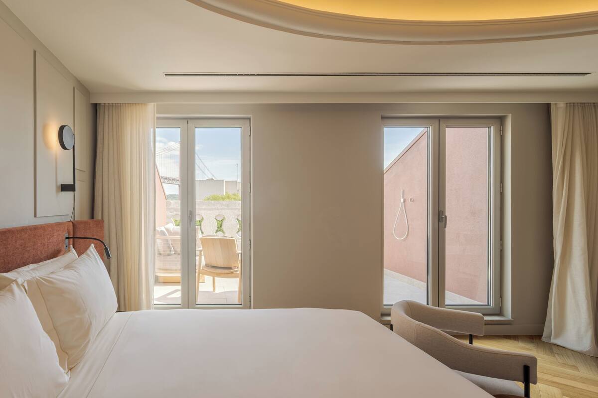 A hotel room with a large bed, a desk, and a chair. There are two windows with curtains, and a balcony is visible through one window.