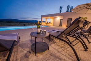 Uma área à beira da piscina com espreguiçadeiras, uma mesa com copos de vinho e um edifício com amplas janelas.