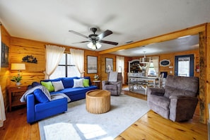 Interior acogedor de una cabaña con un sofá azul, una mesa redonda de madera y dos sillones. Se ve un ventilador de techo y una zona de cocina al fondo.
